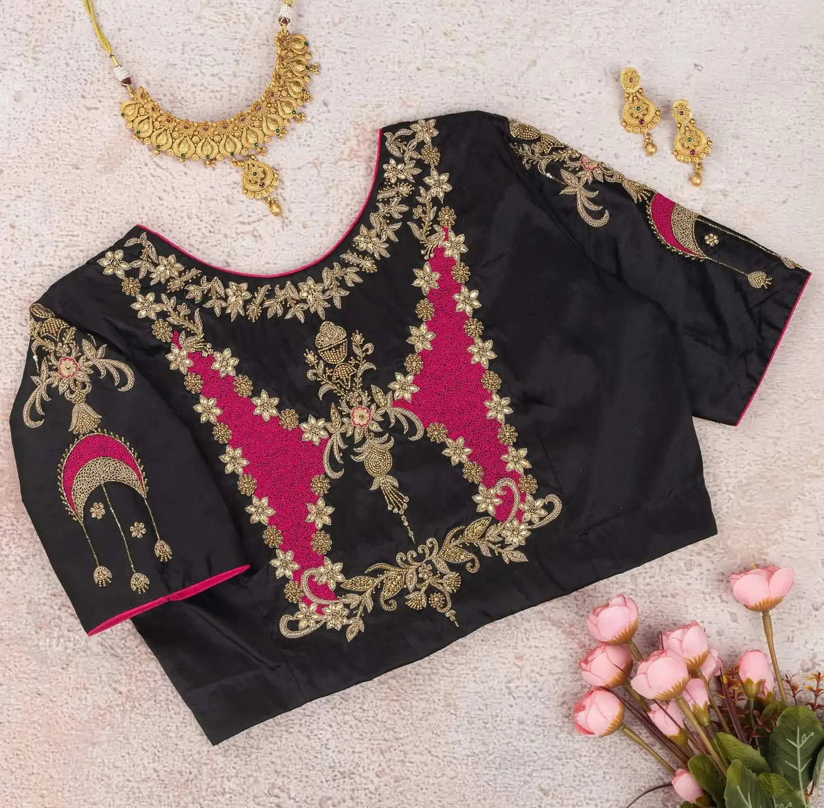 Embroidered back of the black blouse laid flat with gold jewellery and pink roses