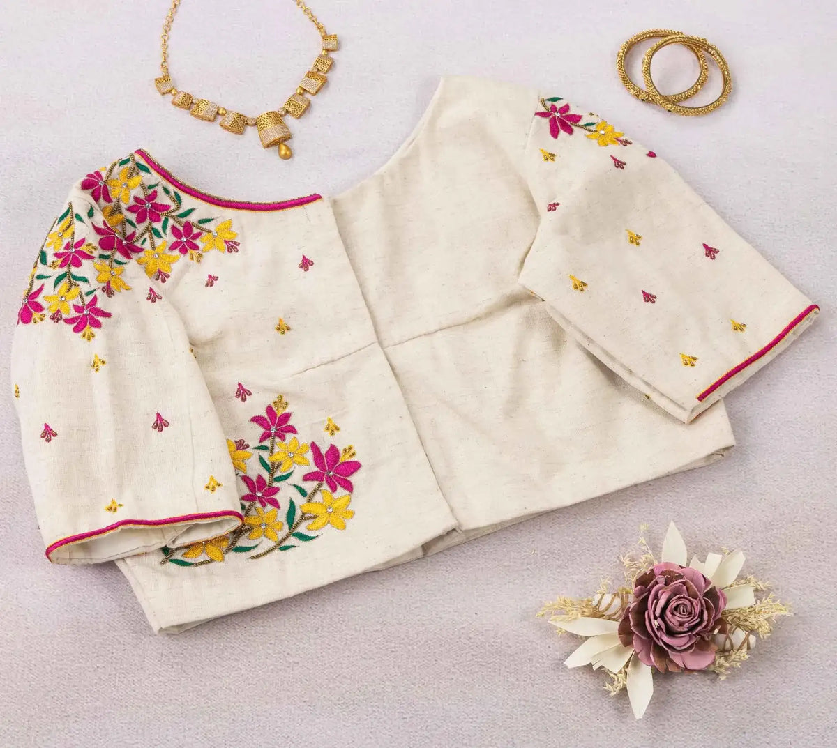 Ivory floral blouse laid out with sleeves spread, gold necklace and bangles alongside