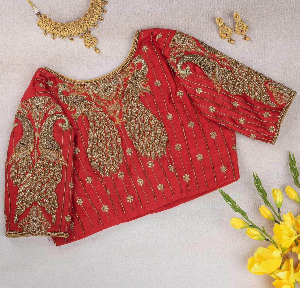 Peacock motif blouse laid flat beside gold jewellery and yellow blooms