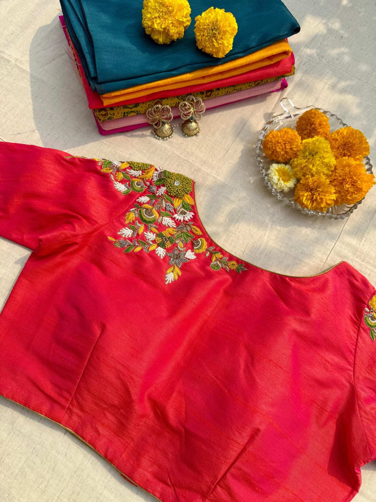 Pink blouse laid flat with floral shoulder embroidery, jhumkas and marigolds alongside
