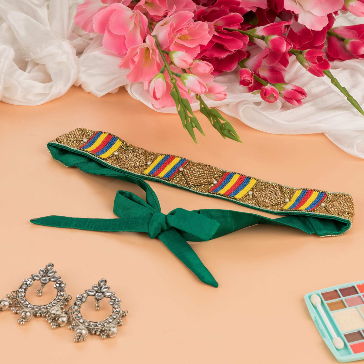 Saree belt with green fabric ties, flat-laid beside silver earrings and pink flowers