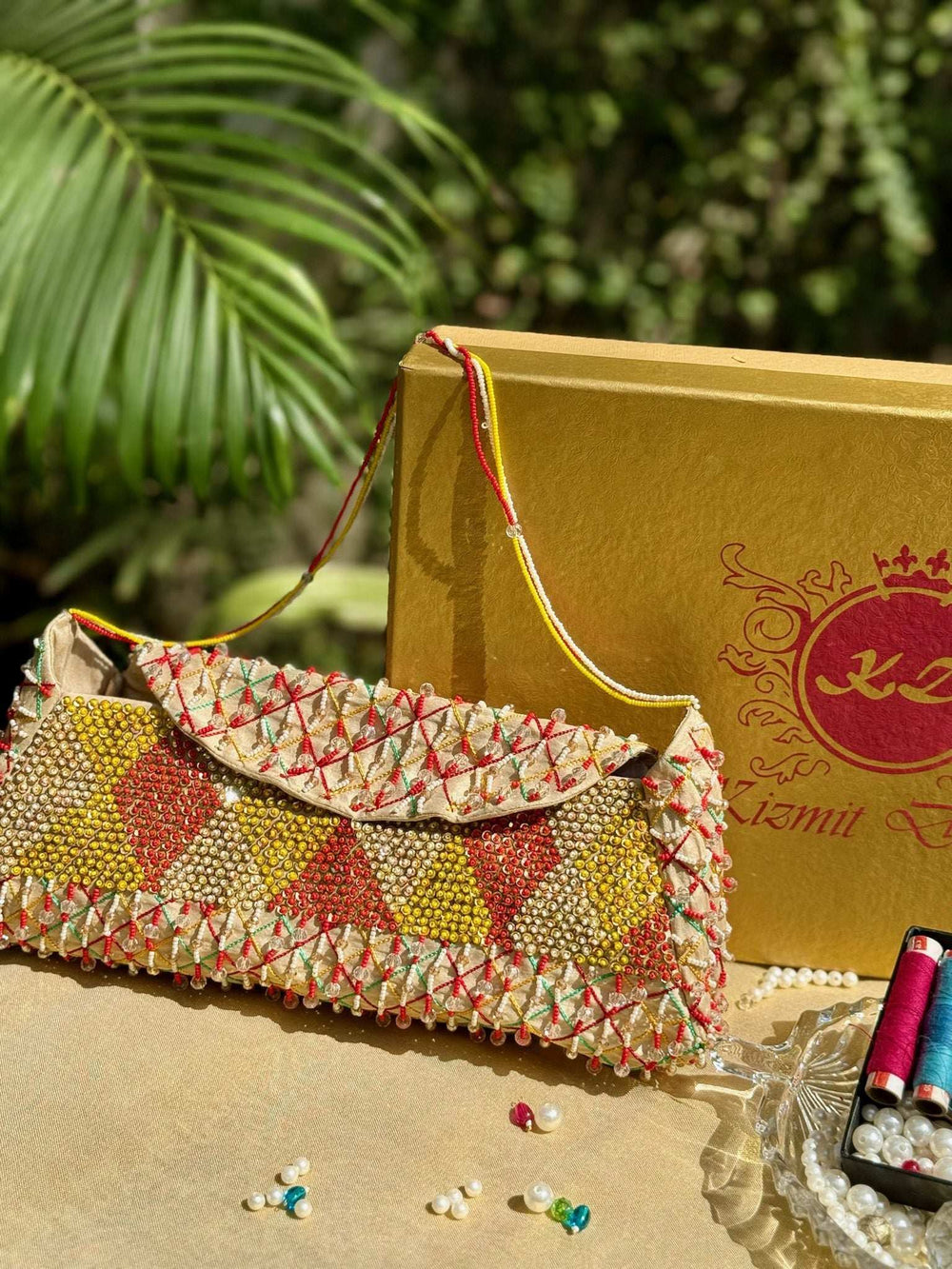 Beaded clutch in red, gold and cream diamonds with a thin bead strap, against a gold gift box