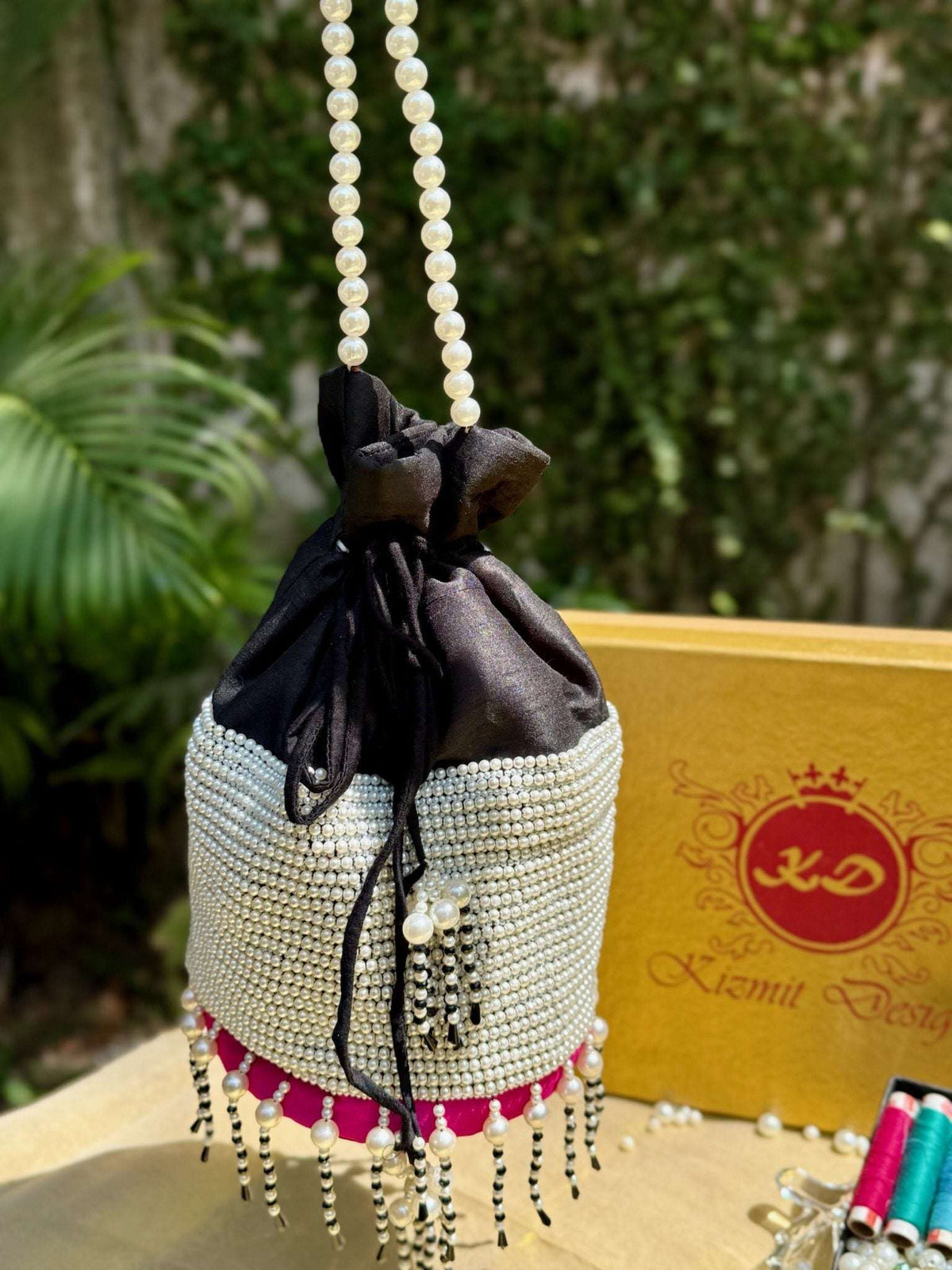 Potli bag suspended by its pearl handle against palm leaves in daylight