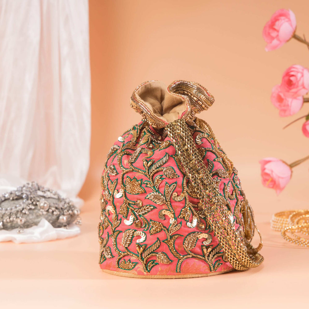 Coral potli bag with gold and green zari floral work, styled beside pink blossoms and silver jewellery