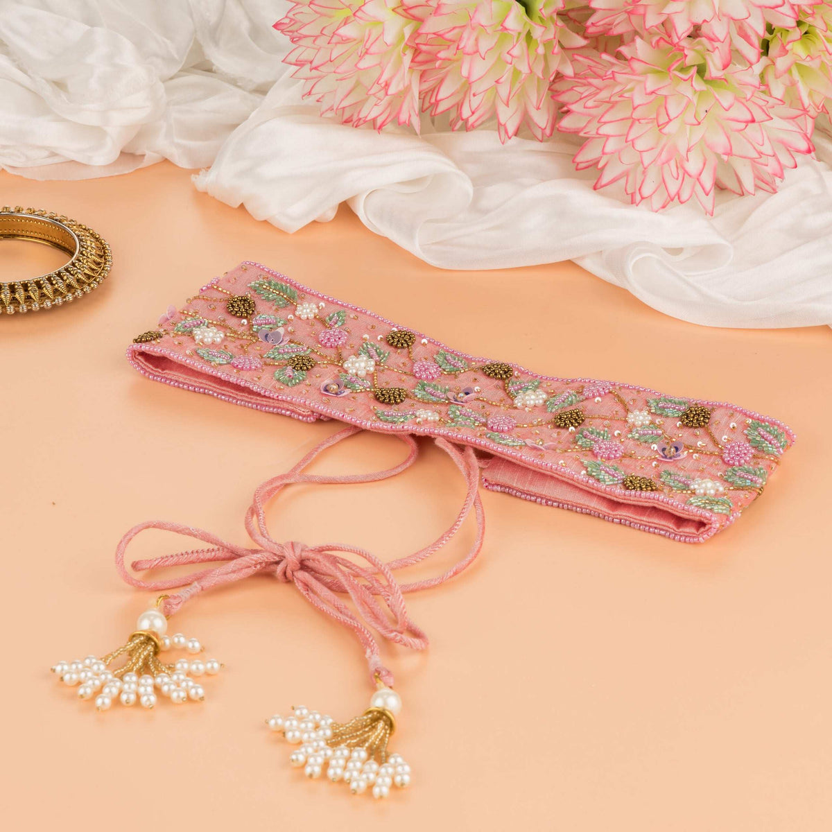 Pink saree belt folded with its cords tied in a bow, pearl tassels and fresh blooms alongside