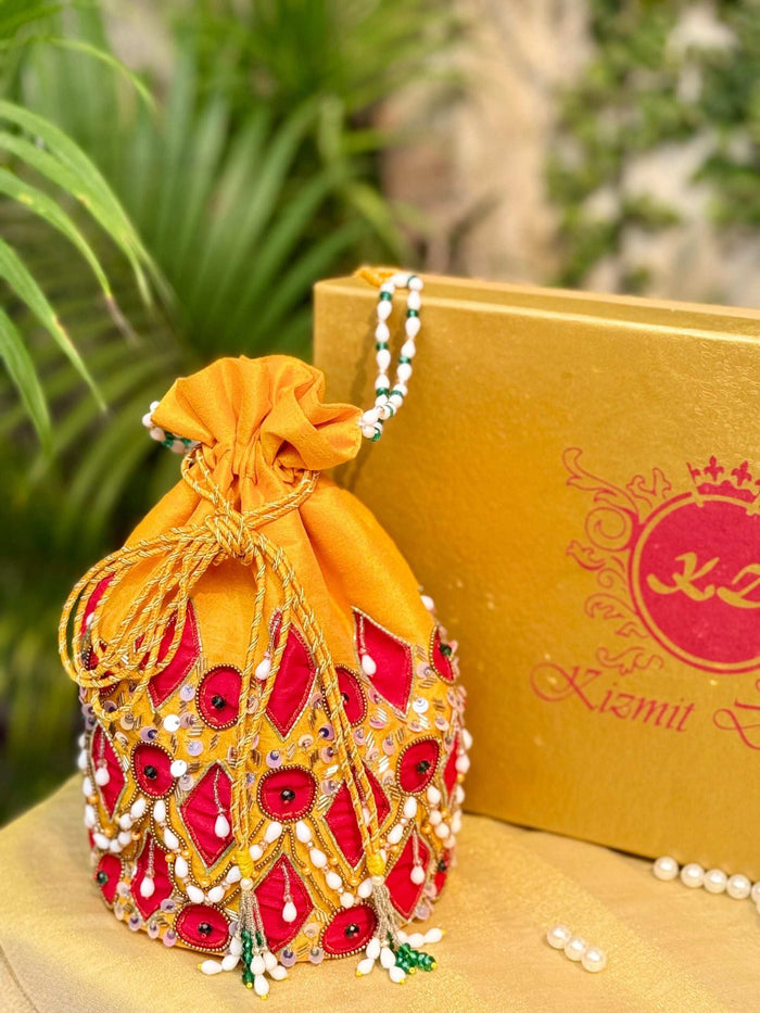Hand embroidered yellow potli with red diamond patches and pearl drops, among leaves