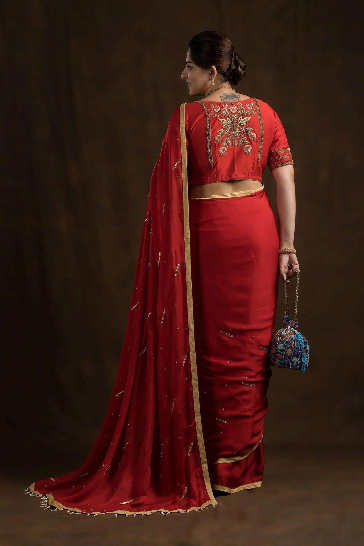 Model seen from behind in a red saree, embroidered blouse back above a gold-edged pallu trailing to the floor