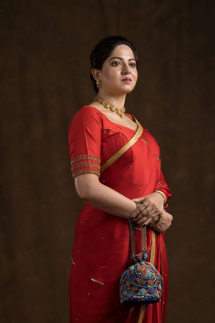 Red blouse with banded gold sleeve worn under a red saree, side view with a blue potli in hand