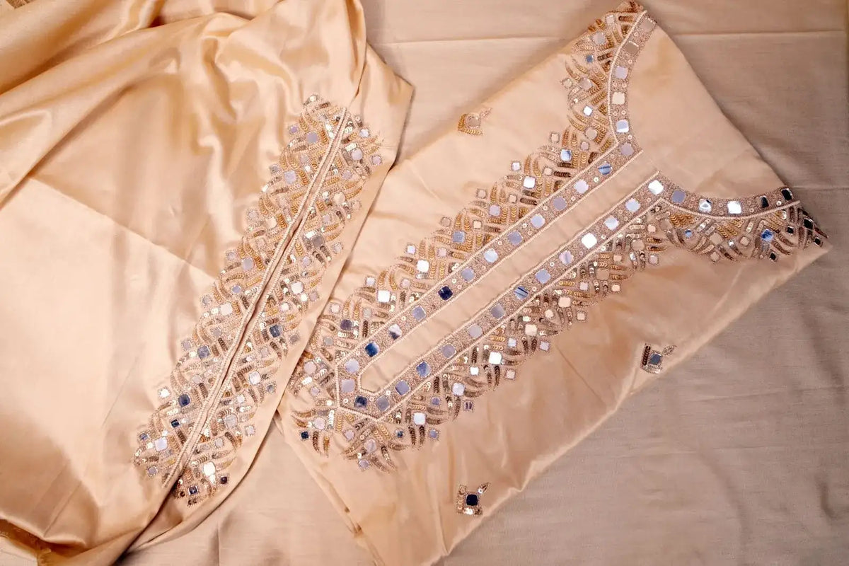 Unstitched beige suit fabric laid flat, mirror-work neckline panel beside the dupatta border