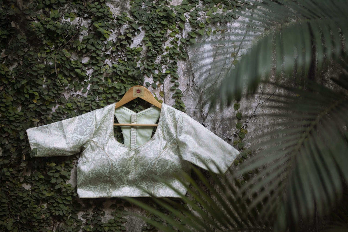 Pistachio green blouse on a hanger against a mossy wall, framed by a palm frond