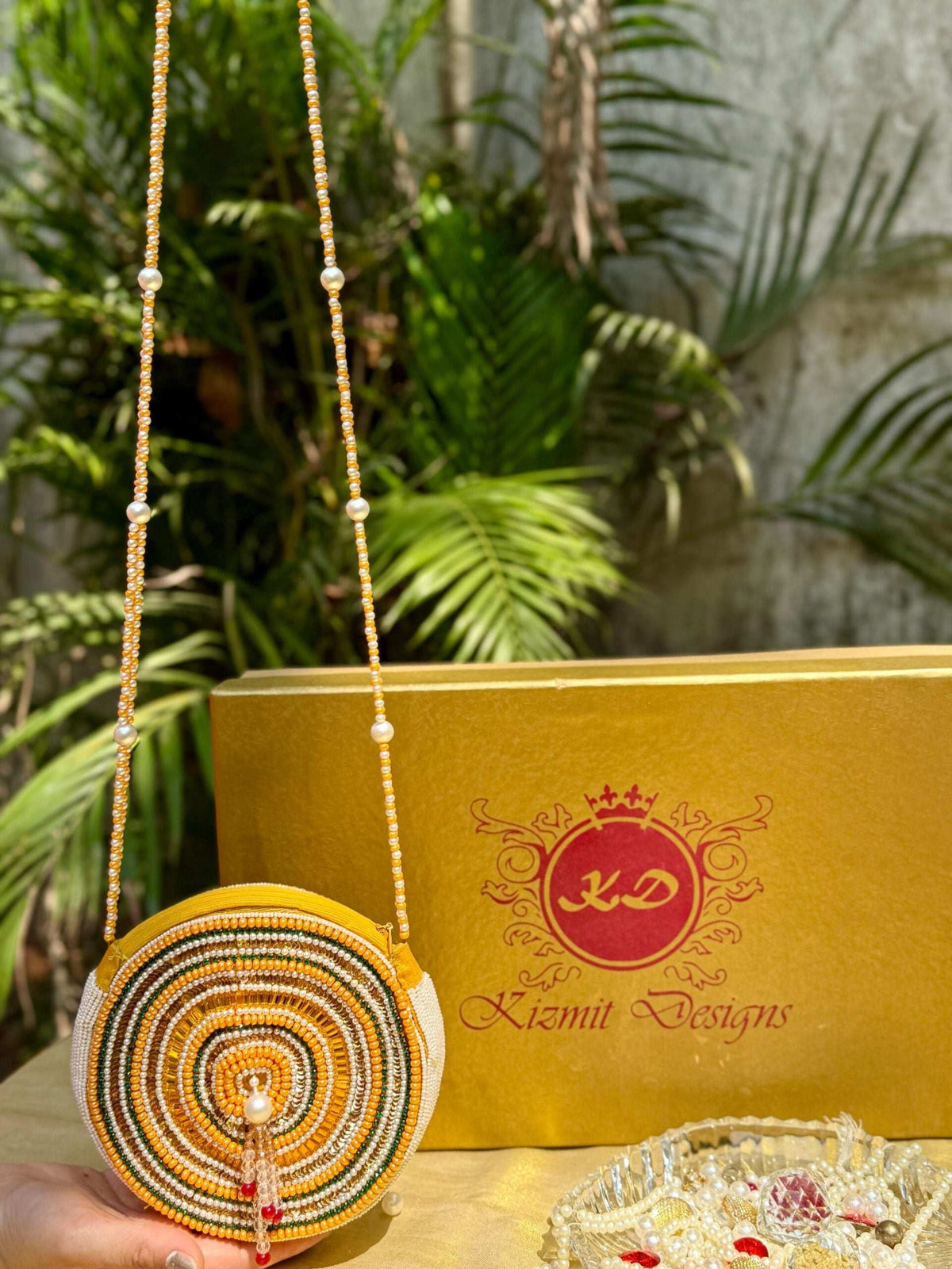 Hand-embroidered round bag with its full-length beaded sling laid out beside a gold box