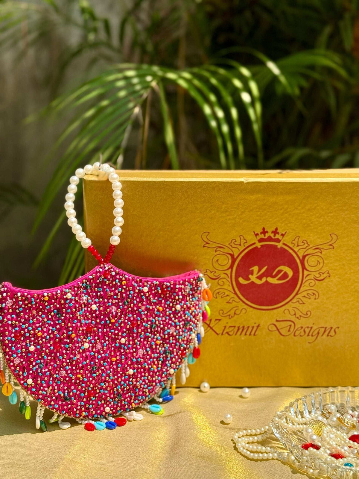 Pink half-moon jhumka clutch in multicolour beads, propped against a gold Kizmit Designs box