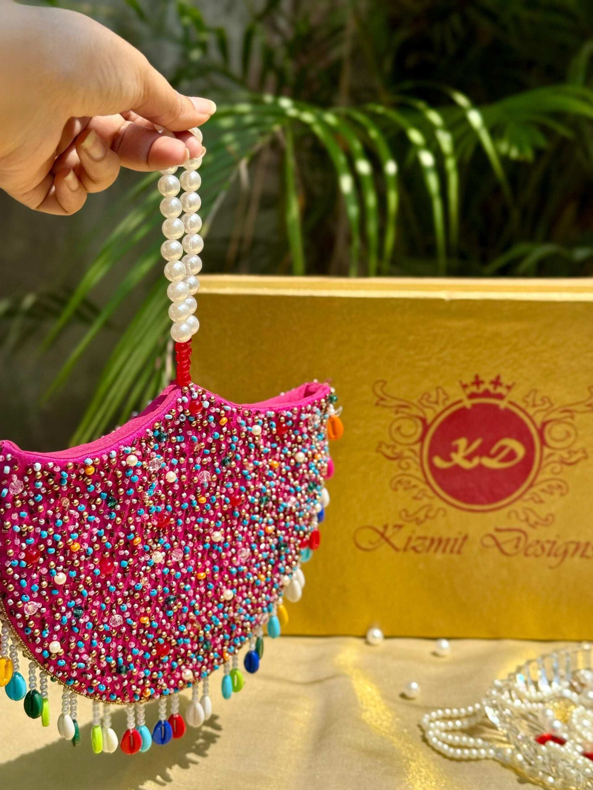 Pearl handle and multicolour bead fringe on the pink jhumka clutch, held beside the gift box