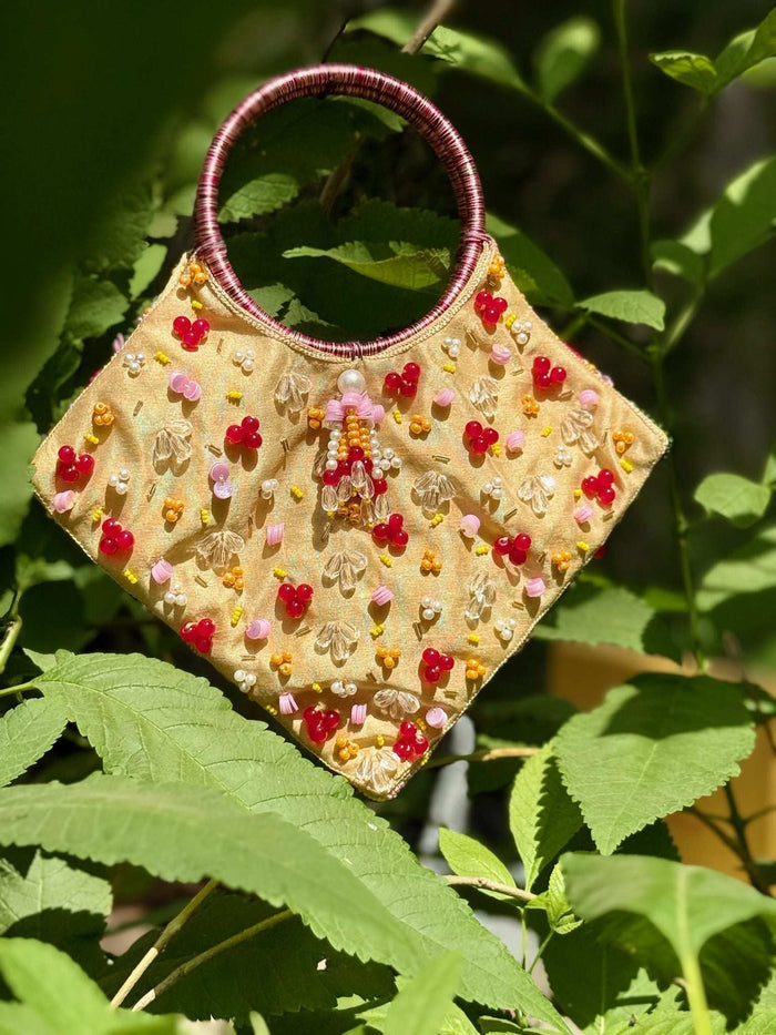 Beige beaded clutch with red and pink flower motifs, hung among sunlit green leaves